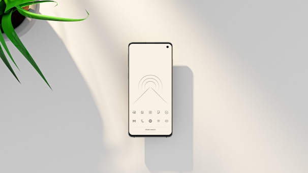 A modern smartphone with a minimalist black and white home screen design sits on a smooth, light-colored surface. Near the top left is part of a green potted plant, adding a touch of nature to the composition.