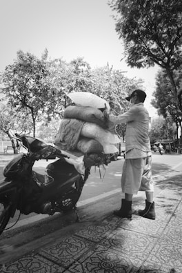 A person wearing a cap and mask stands next to a motorcycle, securing large bags onto the back using ropes. The motorcycle is parked on a decorative tiled sidewalk beside a road. Trees with dense foliage line the street, casting shadows on the pavement.