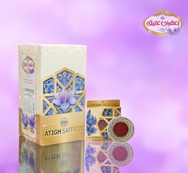 A saffron packaging design featuring a large rectangular box and a smaller round container. The background is a soft gradient of pinks and purples. The packaging is adorned with floral imagery and intricate geometric patterns in shades of blue and gold. The brand name 'ATIGH SAFFRON' is prominently displayed along with a decorative logo.