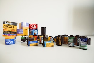 A collection of various film canisters and boxes arranged on a surface. The assortment includes brands such as Kodak, Fuji, and Ilford, with a mix of both color and black-and-white film types. Some canisters are standing upright while others are lying flat, all displayed in a visually appealing way.