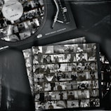 A vintage-style image features a vinyl record player with a record labeled 'Ricky Rique - Love Gun' on the turntable. Nearby, there are film contact sheets displaying black-and-white images of people, including individuals in suits and hats, evoking a classic or retro feel.
