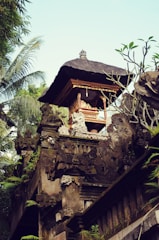 An intricately designed structure with detailed carvings and a thatched roof is surrounded by lush green foliage. The stone structure exhibits ornate decorations in a traditional architectural style, featuring motifs and sculptures.