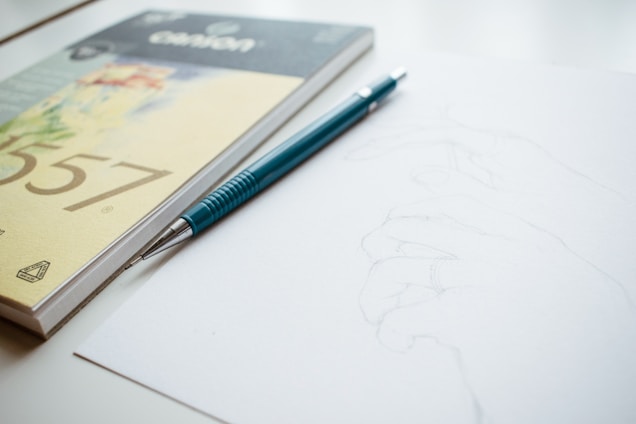 A close-up of an artist's hand sketching delicate graphite lines on textured paper in a softly lit studio.