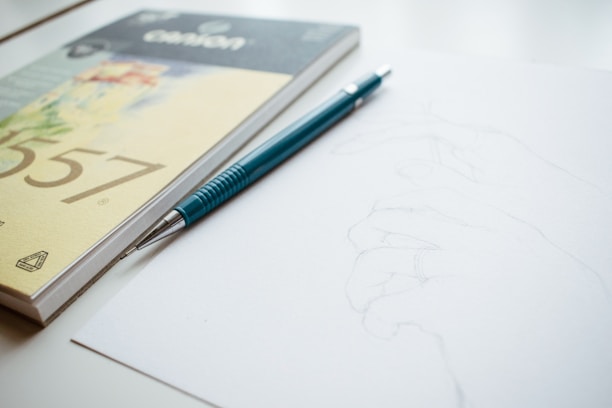 A close-up of a hand holding a sleek, unsharpened pencil over a sketchbook filled with artistic doodles.