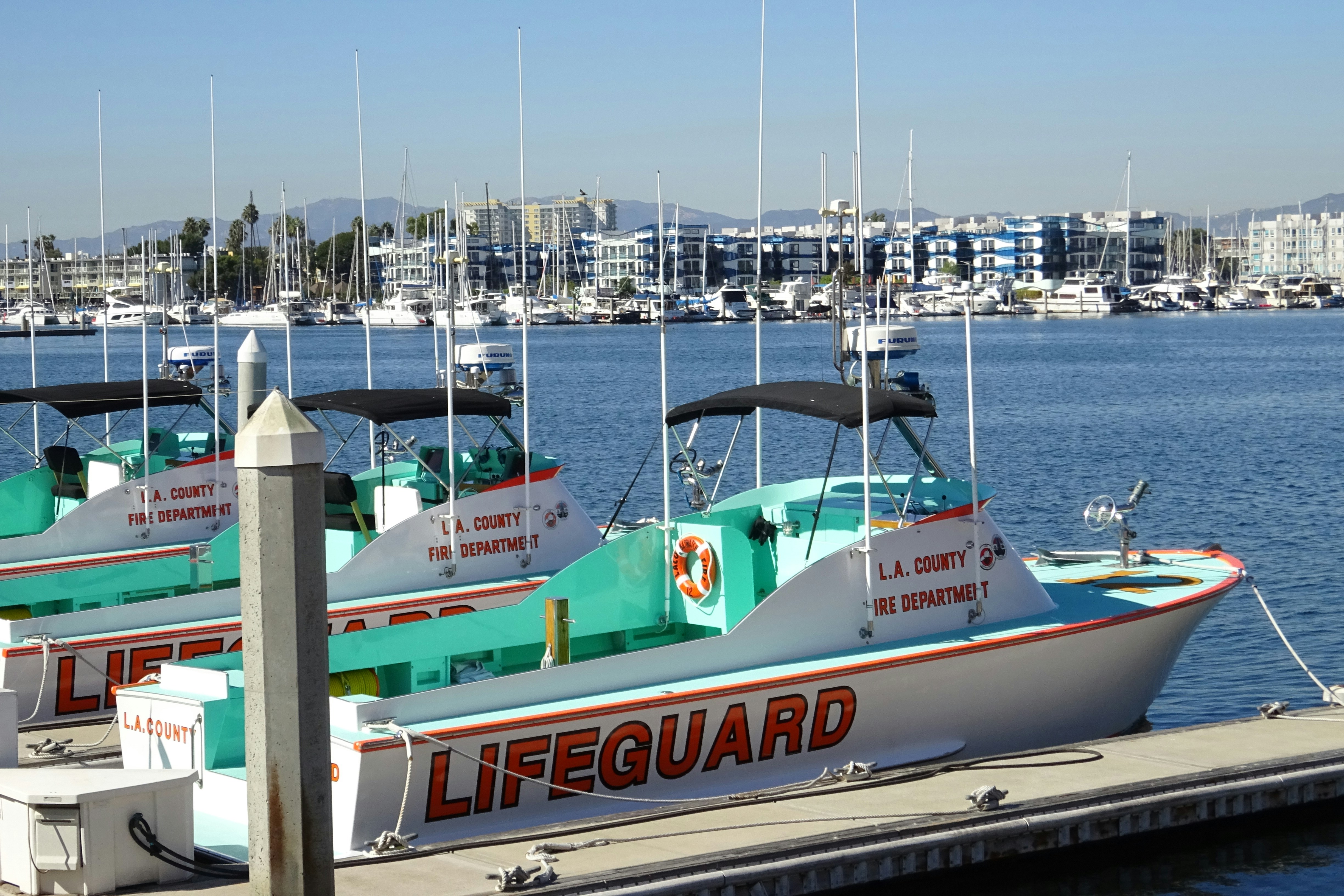 Lifeguard boats docked along a marina with calm water and a cityscape in the background.