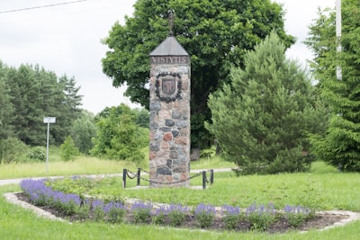 A stone monument stands in a landscaped garden, surrounded by lush green trees and a meticulously maintained flower bed with vibrant purple flowers. The monument features the word 'Vistytis' and a coat of arms or emblem.