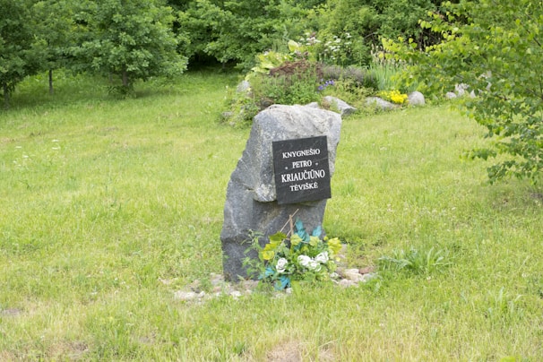 A stone monument stands amidst a lush, green grassy area, surrounded by vibrant trees and various flowering plants. The monument has an engraved plaque with text on it. At the base, there are decorative elements such as white and blue artificial flowers.