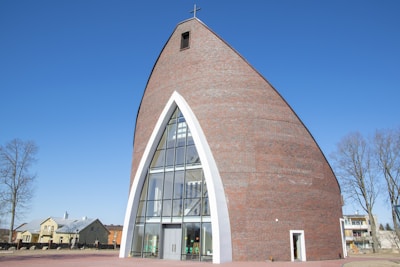 A modern church with a unique, curved architectural design, featuring a large arched glass entrance and a cross on top. The structure is made of brick with white accents framing the entrance. There are a few trees and residential buildings around it, set against a clear blue sky.