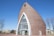 A modern church with a unique, curved architectural design, featuring a large arched glass entrance and a cross on top. The structure is made of brick with white accents framing the entrance. There are a few trees and residential buildings around it, set against a clear blue sky.