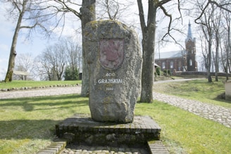 A large stone monument stands in a park setting, inscribed with '400 Gražiškiai 2002' and a coat of arms. The area is surrounded by bare trees and a cobblestone path. In the background, a red-brick church with a tall spire is visible.