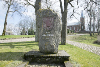 A large stone monument stands in a park setting, inscribed with '400 Gražiškiai 2002' and a coat of arms. The area is surrounded by bare trees and a cobblestone path. In the background, a red-brick church with a tall spire is visible.