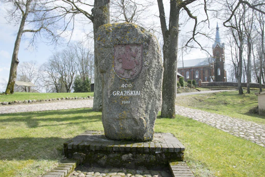 A large stone monument stands in a park setting, inscribed with '400 Gražiškiai 2002' and a coat of arms. The area is surrounded by bare trees and a cobblestone path. In the background, a red-brick church with a tall spire is visible.