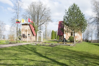 A serene outdoor setting with a small brick structure featuring red roofs and European flags flapping in the breeze. The scene includes a well-maintained grassy area with various trees and shrubs. A white archway and a statue are present, adding to the tranquil atmosphere.