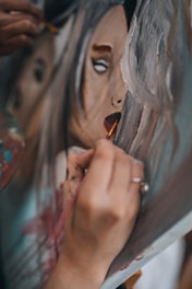 A hand is holding a brush, carefully painting the details on a portrait. The image captures the close-up of the hand in the act of painting, focusing on the delicate brush strokes being added to the lips and face of the portrait. The artwork appears to depict an expressive face with abstract and loose brushwork.