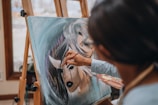 A person is actively painting on a canvas positioned on an easel. The artwork features the face of a woman with dark lipstick and flowing hair, with the painter's hand applying details using a brush. The setting appears to be indoors, with natural light coming through a window.