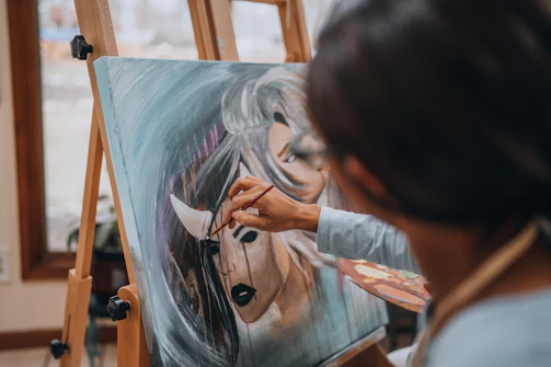 A person is actively painting on a canvas positioned on an easel. The artwork features the face of a woman with dark lipstick and flowing hair, with the painter's hand applying details using a brush. The setting appears to be indoors, with natural light coming through a window.
