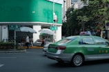 A green and silver taxi is driving on a city street next to a building with green signage at the top. Several people are sitting and standing under large white umbrellas near the corner of the building. There is a prominent exit or pedestrian sign on the wall near the umbrellas. Trees line the street, and tall buildings are visible in the background.
