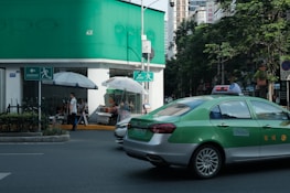 A green and silver taxi is driving on a city street next to a building with green signage at the top. Several people are sitting and standing under large white umbrellas near the corner of the building. There is a prominent exit or pedestrian sign on the wall near the umbrellas. Trees line the street, and tall buildings are visible in the background.