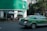 A green and silver taxi is driving on a city street next to a building with green signage at the top. Several people are sitting and standing under large white umbrellas near the corner of the building. There is a prominent exit or pedestrian sign on the wall near the umbrellas. Trees line the street, and tall buildings are visible in the background.
