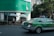 A green and silver taxi is driving on a city street next to a building with green signage at the top. Several people are sitting and standing under large white umbrellas near the corner of the building. There is a prominent exit or pedestrian sign on the wall near the umbrellas. Trees line the street, and tall buildings are visible in the background.