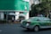 A green and silver taxi is driving on a city street next to a building with green signage at the top. Several people are sitting and standing under large white umbrellas near the corner of the building. There is a prominent exit or pedestrian sign on the wall near the umbrellas. Trees line the street, and tall buildings are visible in the background.