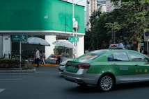 A green and silver taxi is driving on a city street next to a building with green signage at the top. Several people are sitting and standing under large white umbrellas near the corner of the building. There is a prominent exit or pedestrian sign on the wall near the umbrellas. Trees line the street, and tall buildings are visible in the background.