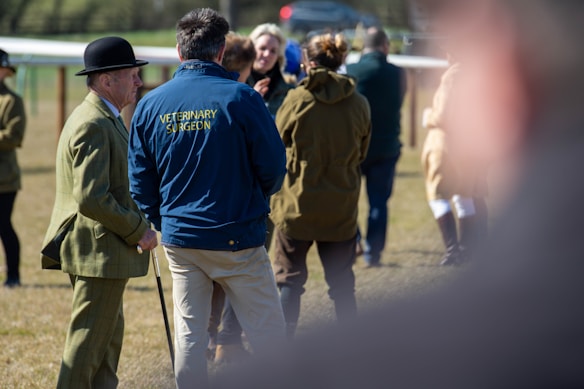 Several people are gathered outdoors on a sunny day. One man is wearing a green suit and bowler hat while another person in a blue jacket has 'Veterinary Surgeon' written on the back. Others are dressed in casual and outdoor clothing, possibly indicating a countryside or equestrian setting.