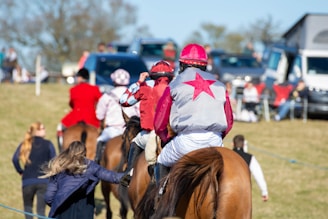 A group of people mounted on horses, wearing colorful jockey outfits. They are riding across a grassy field. In the background, there are more people and parked vehicles, indicating a social or sporting event. A woman in the foreground, on foot, appears to be interacting with the riders.