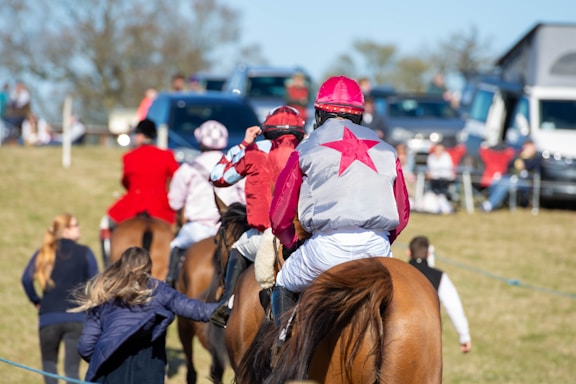 A group of people mounted on horses, wearing colorful jockey outfits. They are riding across a grassy field. In the background, there are more people and parked vehicles, indicating a social or sporting event. A woman in the foreground, on foot, appears to be interacting with the riders.