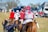 A group of people mounted on horses, wearing colorful jockey outfits. They are riding across a grassy field. In the background, there are more people and parked vehicles, indicating a social or sporting event. A woman in the foreground, on foot, appears to be interacting with the riders.