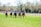 A group of jockeys are riding horses at full speed across a grassy field, with trees and a fence in the background. The jockeys are wearing colorful racing attire, including helmets and eye coverings, while the horses are equipped with saddles and bridles.