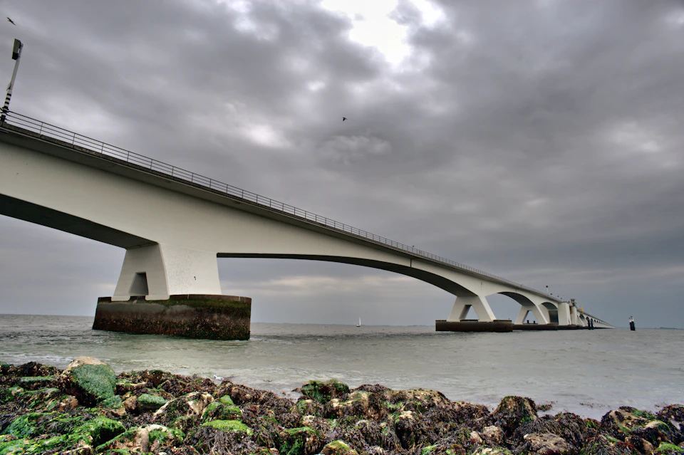 White bridge with two arches spans across ocean, supported by concrete pillars in foreground