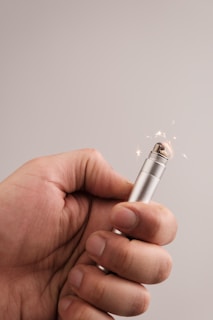 A hand holding a metal lighter with a flame visible at the top and small sparks flying around.