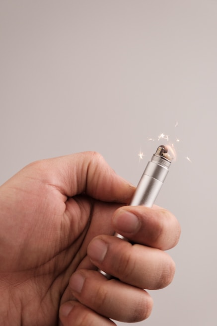 A hand holding a metal lighter with a flame visible at the top and small sparks flying around.