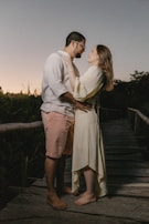 A couple stands closely on a wooden pathway surrounded by nature. The man wears a light shirt and shorts, while the woman is in a flowing dress. They share an affectionate gaze as the sun sets, casting a soft glow over the scene.