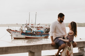 A fishing boat named 'Menino do Rio' is moored on calm waters. The boat's hull is painted white and blue with red accents. In the foreground, a man and a woman are sitting on a railing, slightly out of focus, suggesting an intimate or romantic moment. The background is serene, with the water extending to the horizon and vague outlines of trees or other landforms.
