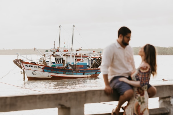 A fishing boat named 'Menino do Rio' is moored on calm waters. The boat's hull is painted white and blue with red accents. In the foreground, a man and a woman are sitting on a railing, slightly out of focus, suggesting an intimate or romantic moment. The background is serene, with the water extending to the horizon and vague outlines of trees or other landforms.
