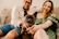 A happy couple cuddling their playful fluffy French bulldog puppy in a sunlit living room.