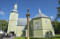 A large green wooden church with a metallic dome and tower is set against a bright blue sky. A group of people gathers near the entrance, and a wooden cross stands in the foreground. The surrounding trees are lush and green, accentuating the peaceful atmosphere.