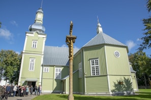 A large green wooden church with a metallic dome and tower is set against a bright blue sky. A group of people gathers near the entrance, and a wooden cross stands in the foreground. The surrounding trees are lush and green, accentuating the peaceful atmosphere.