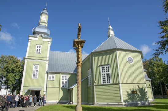 A large green wooden church with a metallic dome and tower is set against a bright blue sky. A group of people gathers near the entrance, and a wooden cross stands in the foreground. The surrounding trees are lush and green, accentuating the peaceful atmosphere.