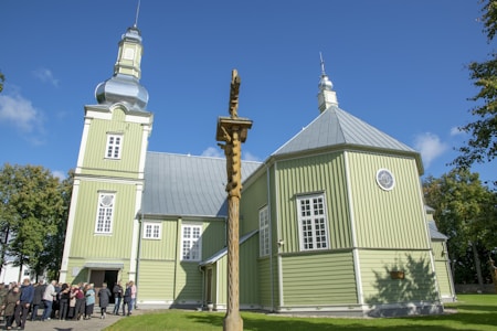 A large green wooden church with a metallic dome and tower is set against a bright blue sky. A group of people gathers near the entrance, and a wooden cross stands in the foreground. The surrounding trees are lush and green, accentuating the peaceful atmosphere.