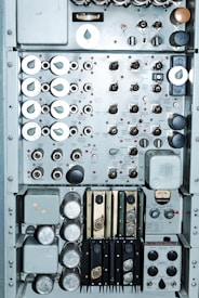 A close-up view of an intricate control panel featuring numerous dials, switches, and gauges. The panel includes labeled components with a mix of metallic knobs and buttons. Various measurement instruments and inputs are visible, possibly indicating functionality related to electronic or mechanical operations. The arrangement suggests a complex system, possibly from a vintage or industrial setting.