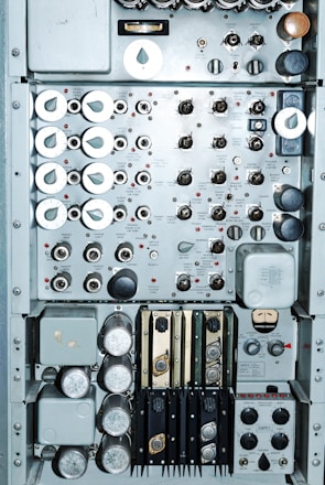 A close-up view of an intricate control panel featuring numerous dials, switches, and gauges. The panel includes labeled components with a mix of metallic knobs and buttons. Various measurement instruments and inputs are visible, possibly indicating functionality related to electronic or mechanical operations. The arrangement suggests a complex system, possibly from a vintage or industrial setting.
