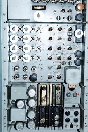 A close-up view of an intricate control panel featuring numerous dials, switches, and gauges. The panel includes labeled components with a mix of metallic knobs and buttons. Various measurement instruments and inputs are visible, possibly indicating functionality related to electronic or mechanical operations. The arrangement suggests a complex system, possibly from a vintage or industrial setting.