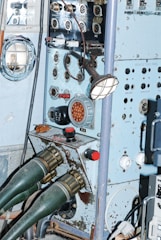 A vintage control panel with various dials, switches, and connectors. The panel is light blue with signs of rust and aging. Two large green connectors are attached to the lower part of the panel. It features a combination of metal and plastic components, with a grid-protected light on the side.