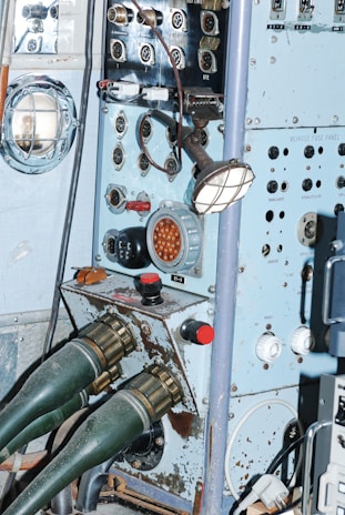 A vintage control panel with various dials, switches, and connectors. The panel is light blue with signs of rust and aging. Two large green connectors are attached to the lower part of the panel. It features a combination of metal and plastic components, with a grid-protected light on the side.