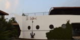 The image features a building facade designed to resemble the front of a cruise ship. It includes elements such as round windows and a large decorative anchor attached to the white wall. The words 'S.S. Safari' are painted on the side. The structure is surrounded by lush greenery and palm trees.