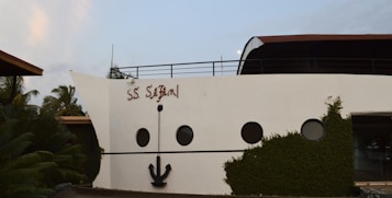 The image features a building facade designed to resemble the front of a cruise ship. It includes elements such as round windows and a large decorative anchor attached to the white wall. The words 'S.S. Safari' are painted on the side. The structure is surrounded by lush greenery and palm trees.