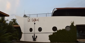 The image features a building facade designed to resemble the front of a cruise ship. It includes elements such as round windows and a large decorative anchor attached to the white wall. The words 'S.S. Safari' are painted on the side. The structure is surrounded by lush greenery and palm trees.
